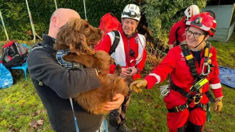 SARA Rescue A brown spaniel is being carried by a man while firefighters in red uniform stand around them smiling.