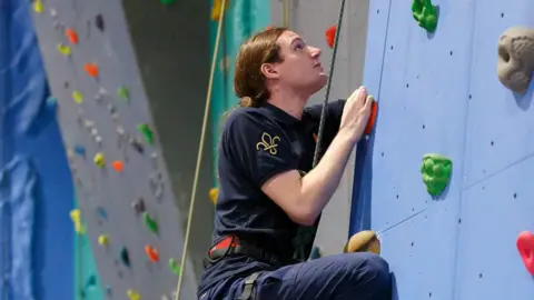 Sunderland District Scouts Aaron Horton photographed at an indoor climbing competition. He is wearing navy blue trousers and t-shirt and is rock climbing. His brown hair is tied at the back.