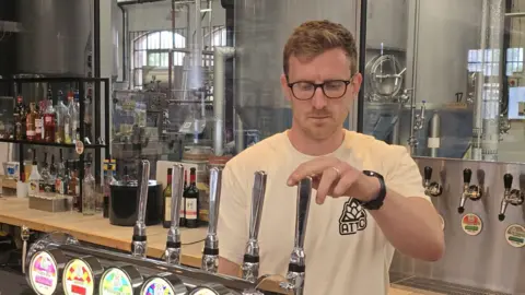 A man with blond hair stands behind a bar with his hand on a beer pump.