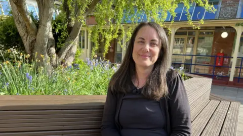Helen is pictured sitting on a bench. She is wearing a black cardigan and gold necklace and is gently smiling at the camera. Behind her, there are spring flowers and a tree with a building in the background which has lots of windows. 