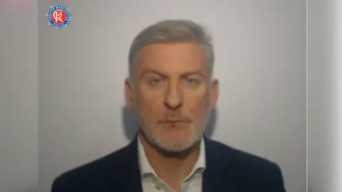 GMP Man with blue eyes and grey hair wears a suit and blazer in a mugshot.