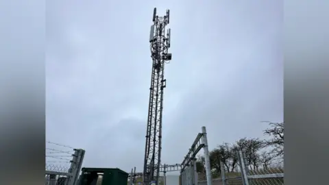 Three and Beacon Comms The mast tower with fencing around it on a cloudy day.