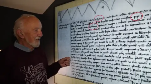 Shaun Whitmore/BBC A man with grey hair and a beard standing by a screen, on which is Old English handwriting