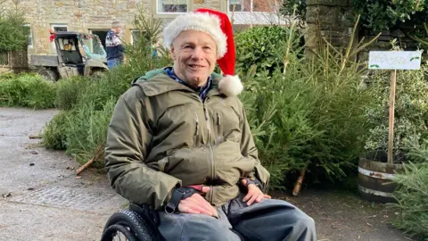 Rob wearing a Santa hat and a green winter jacket is seated in a wheelchair on an outdoor path. Behind him is several evergreen trees are arranged for sale, and a small wooden sign is visible near a stone wall.