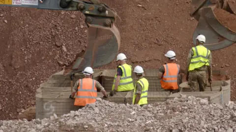 Five people, two in orange hi-vis vests and three in yellow ones, stand around a barrier constructed around an unseen 50kg bomb discovered in Coventry, while two diggers cover the device with sand to minimise damage during an explosion.