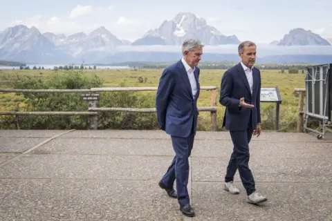 Getty Images Jerome Powell, presidente do Federal Reserve dos EUA (à esquerda) e Mark Carney, ex -governador do Banco da Inglaterra, andam pelos jardins do Simpósio Econômico de Jackson Hole em 2019. Ambos vestindo ternos azuis, estão cercados por campos verdes exuberantes com as montanhas ao fundo