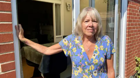 Una mujer rubia con un vestido azul floreado está de pie frente a la puerta del patio de un bungalow.