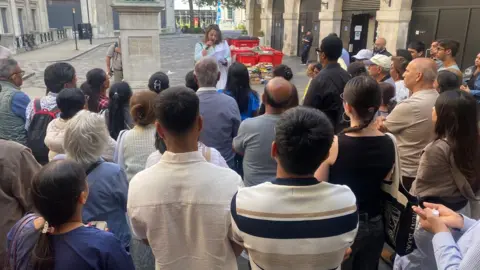 Vigil outside the Indian Embassy where a crowd has gathered and a woman in front is speaking into a microphone