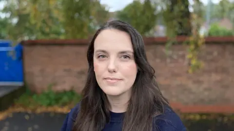 Luke Deal/BBC Bex Patch with long dark brown hair and a navy jumper staring into the camera. A brick wall is in the background. 