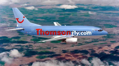 PA Media A passenger plane branded with Thomsonfly on the side flying in the air with light clouds surrounding it. The ground below appears bare without any buildings.