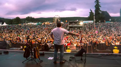 Hope Simmers Cammy Barnes, with his back to the camera, surveys the crowd at Belladrum's main stage on Saturday night. People are holding up phones with the torches on.