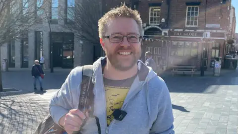 BBC/Tom McDougall A man with light hair and a beard wearing a grey hoodie squints in the sunshine