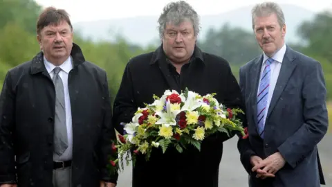 Pacemaker Three men standing side by side as one holds a wreath. From left to right is a man with short brown hair black coat, grey trousers, white shirt and grey tie. then a man with short grey hair and a black coat. Then a man with short grey hair, grey moustache wearing grey jacket, white shirt, blue striped tie and black trousers. Behind them is a road where the attack occured.