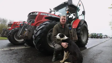 PA Media An older man with short greying hair wearing an olive green fleece, work trousers and wellies. He is kneeling down in front of a red tractor, and is holding a black and white collie dog. 