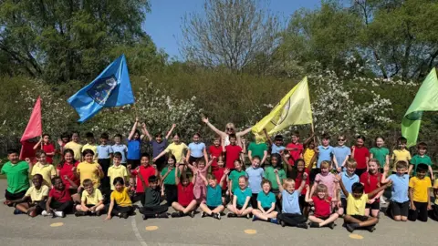Swindon Borough Council A large group of schoolchildren wearing black shorts and skirts with colourful red, green, blue and yellow t-shirts. They are sitting on a playground in front of trees and bushes, holding up large flags which match their shirt colours.