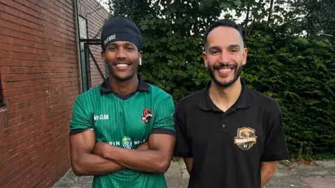 Youcef El-Barhdadi Jabir Abdalla (left) and Youcef El-Barhdadi pose for a photo side-by-side next to a fence and the side of a brick building. Jabir has muscled arms folded and wears a head covering and a green football shirt. Youcef's arms are behind him - he has dark hair shaved at the sides and a beard and wears a black polo shirt.