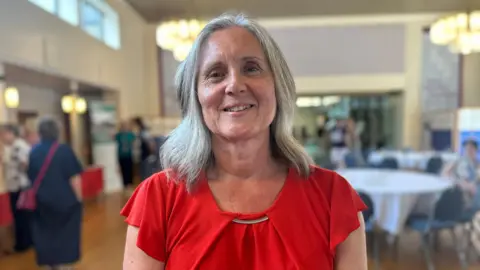 Helen Moss smiles at the camera. She has shoulder-length silver hair and wears a red top. Behind her people are milling around looking at an exhibition