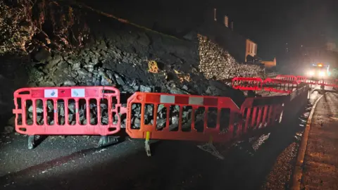 A high-visibility highways barricade cordons off a section of road where a stone wall has collapsed. There is debris from the wall halfway across the road.