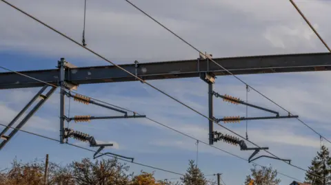 A view of the cables as an electric powered train passes through a British railway station. It is a sunny day. The tips of trees and the apex of a home can be seen. 