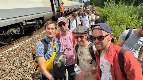 Stornoway gather on the railway sidings. Oli is holding his guitar case. Dozens of passengers are gathered behind them.