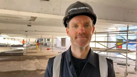 BBC A man stands in a multi-storey car park and smiles, while wearing a black hat with says Capital&Centric. There is construction work taking place behind him.