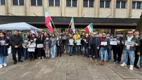 A group of people gathered in Cheltenham Town Centre in front of a big shop building. Some of them have Iranian flags and many are holding signs in their hands.