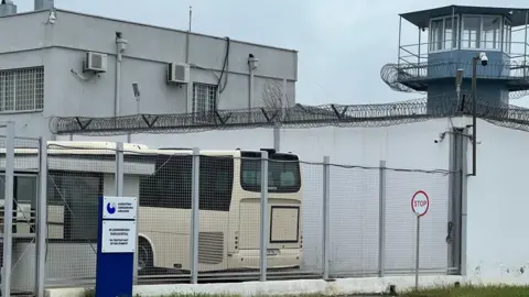 Rayhan Demytrie A white prison entrance with wire fences and barbed wire surrounding a watchtower and the top of the walls.