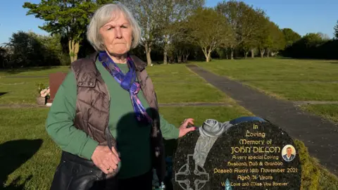 Ann Dillon is wearing a green jumper and a black jacket on top. She is standing next to her husband's grave, marked with a black and gold headstone. She has her left hand resting on it. 
