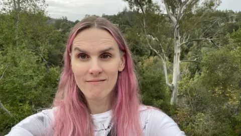 Supplied/Rana Charron An image of a woman with long, pink straight hair smiling at the camera. Behind her is a backdrop of trees. 