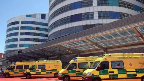 Getty Images The sun is shining outside the A&E department at the Queen Elizabeth Hospital in Birmingham. A line of yellow ambulances is lined up below a large glass awning, above which can be seen two curved glass sections of the hospital itself.