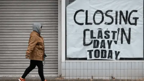 A person walks past a closed down shop which says "Closing down. Last day today" in the window. 