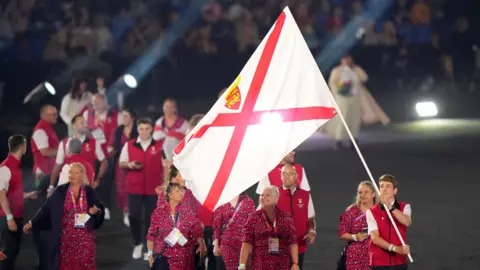 Jersey's 2022 Commonwealth Games team walk out into a stadium during the event's opening ceremony in Birmingham. A man at the front is carrying a Jersey flag.