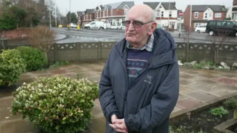 An elderly man stands with his hands clasped in a garden