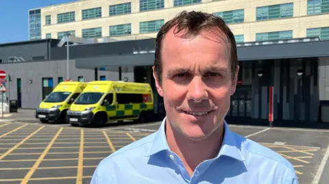 Benny Goodman, chief operating officer, is facing the camera wearing a blue open collar shirt. He is standing in front of the hospital doors which are black frames with long glass windows. Above are the wards with beige brick and glass windows. He is standing on black tarmac in the car park which has yellow cross hatch lines on it. Over his left shoulder are two bright yellow and green ambulances parked outside the entrance to the hospital.