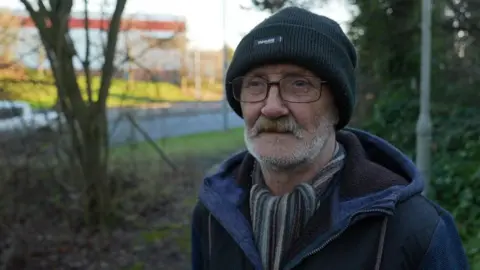 Vince Connor is wearing glasses, a black wooly hat and a striped scarf. Behind him are trees and bushes.