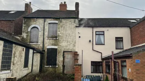 The rear of two terraced homes with the left one covered in green mould and bricked up windows. The home on the right is clean and well-maintained.