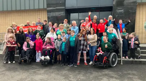 A number of pupils and staff from Ardnashee School & College, as well as members of The Undertones, are outside the movie theatre. Some are waving their hands, while others are holding a Derry City flag.
