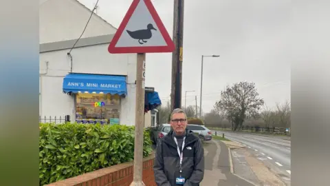 John Knott stands in front of a lamp post with a triangular sign that has a black and white image of a duck. Knott looks unhappy and is wearing a black jacket with a lanyard around his neck. Behind him is a shop with a sign reading "Ann's Mini Market". The are a couple of cars parked in front of the building. 