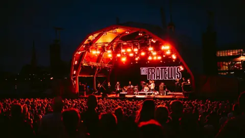Nadine Ballantyne The Fratellis performing at Bristol Sounds. The stage is lit in red and the band is standing in front of their logo, projected on the back in capital letters. There is a huge crowd watching them.