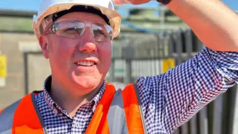 Paul is wearing a white hard hat, blue and white chequered shirt and an orange hi-vis jacket with clear safety goggles. His arm is raised up with his hand shielding the sun from his eyes.