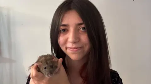 A young woman with long brown hair and a nose ring holds up a rat. She is standing in a room with white walls behind her.