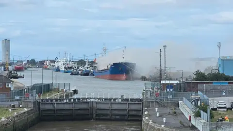 Anthony Smith A view of a dock through the lock gates. A blue and red vessel has grey smoke billowing from its deck. Other ships can be seen docked alongside.