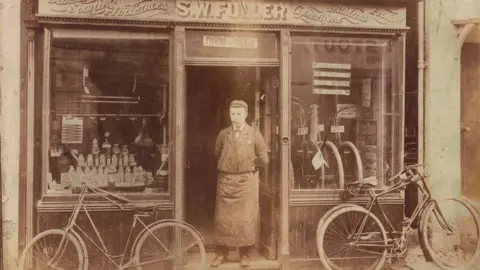 an aged sepia photograph SW Fuller standing outside his shop in about 1892, between two bicycles