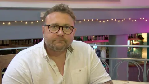 David Jenkins looking at the camera wearing a white t-shirt. He is wearing black glasses and has a short beard. In the background is a string of fairy lights across the wall and a bar below