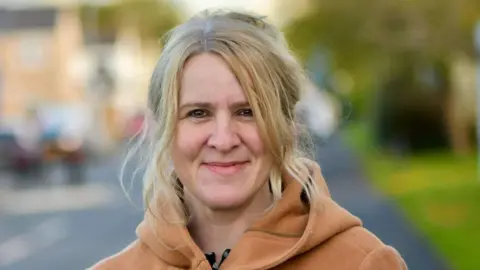 Councillor Ella Billiald a woman with wavy blonde hair half pulled up and an orangey-beige coat with a hood, her background is blurred
