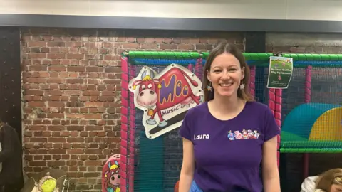 Laura Gilbert, wearing a purple t-shirt, stood in front of colourful children play equipment. There is a brick wall behind the equipment. 