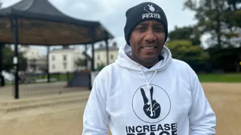 Delroy Ellis is standing in a park. He is wearing a white hoodie with Increase the Peace written on it.