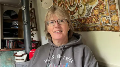 Ally Clarke is sitting inside her canal boat which has cream walls. Behind her is a tapestry, a stove and a kettle. She has medium length blonde hair and is wearing glasses and a grey jumper with 'this is how i roll' printed on the front. 