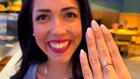 A close up of a young woman smiling broadly and holding her hand up in front of her. She has a ring on her middle finger with a sapphire and diamond.