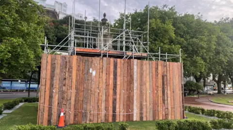 Torbay Council Scaffolding and wooden boards up around Torquay's war memorial. An orange traffic cone is also propped up by the boards and grass and shrubberies surround the memorial.